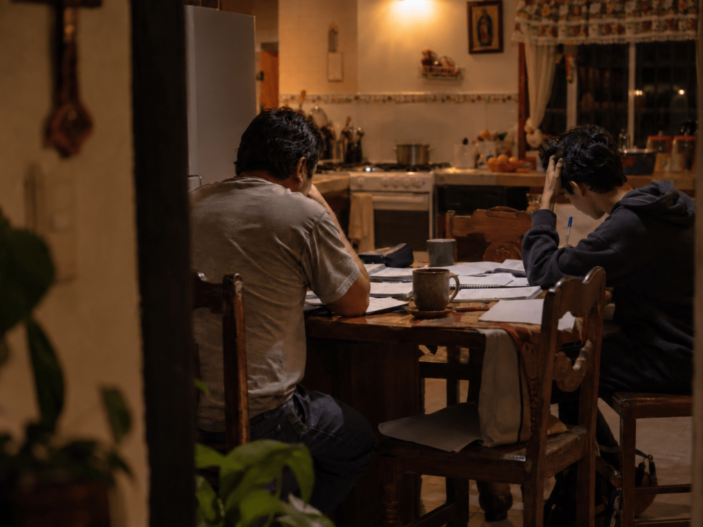 Mesa de cocina de noche con papeles de escuela, donde un papá y su hijo adolescente conversan sin mirarse