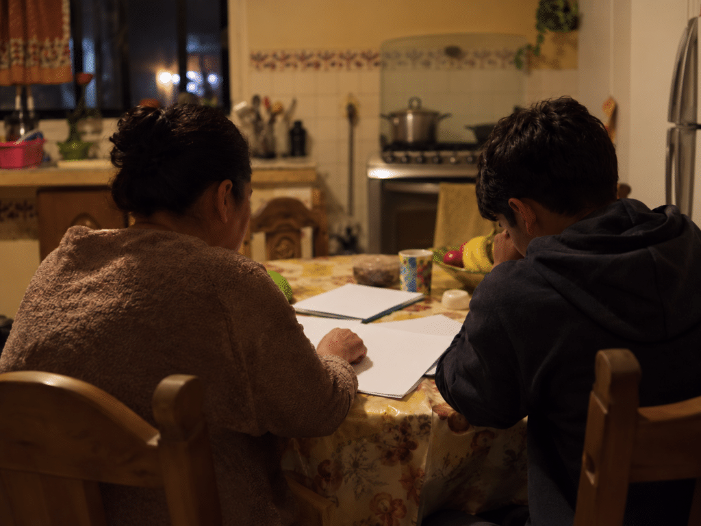 Mesa de cocina de noche con papeles de la escuela extendidos mientras una mamá y su hijo adolescente conversan vistos desde atrás