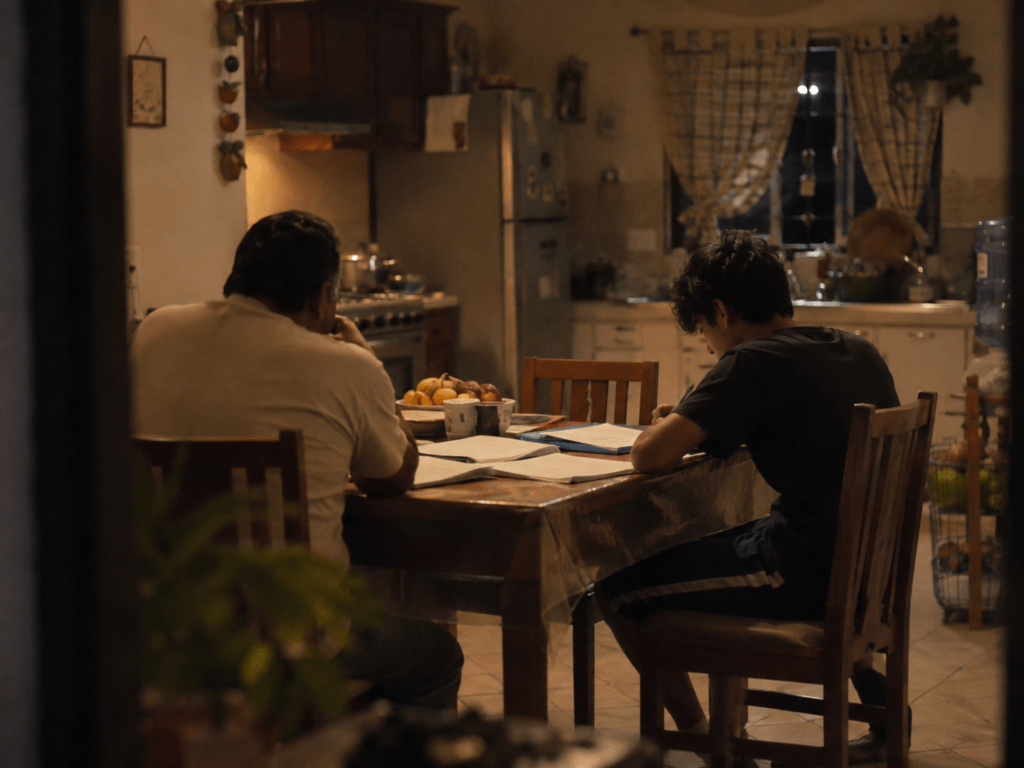 Mesa de cocina de noche con papeles escolares esparcidos mientras un padre y su hijo adolescente conversan sin mirarse