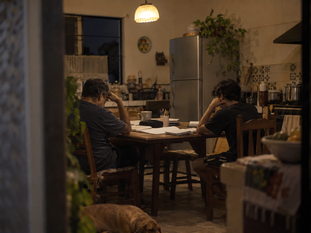 Mesa de cocina de noche con papeles escolares extendidos donde un padre y su hijo adolescente conversan sin mirarse